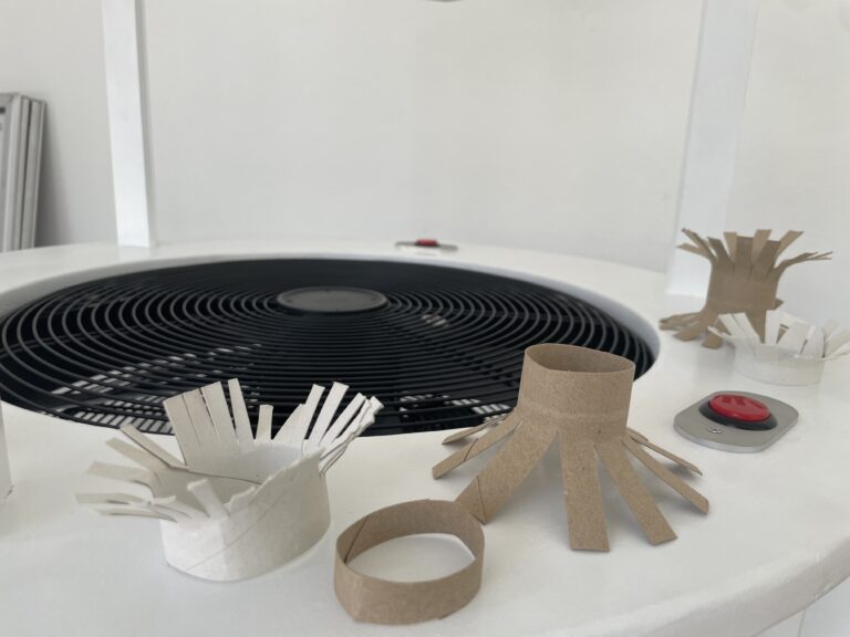 Das Bild zeigt einen Ausschnitt des Windkanals auf dem ein Ventilator zu sehen ist neben dem verschiedenen Flugobjekte aus Papprolle liegen.
